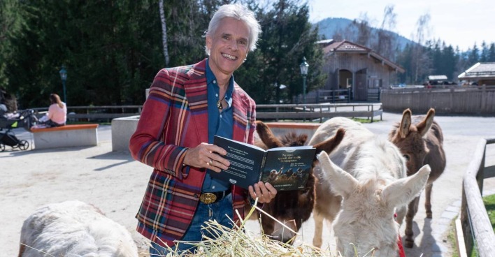 Thomas Brezina mit einem Buch, einem karierten Sakko und umgeben von Eseln.
