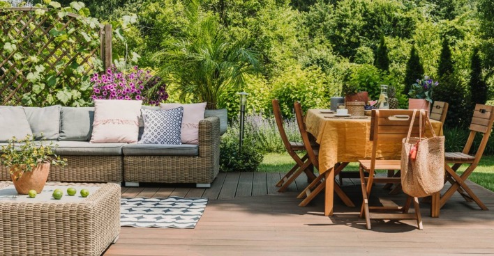 Blick in einen Garten mit Holztisch und Sesseln. Im Hintergrund ein Rattansofa.