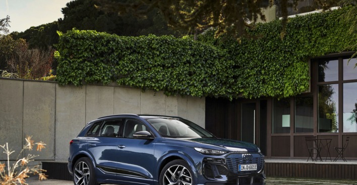 Ein blau glänzender Audi Q6 e-tron steht auf einer Terrasse. Im Hintergrund sind Efeuranken, ein Tisch und Sesseln zu sehen.