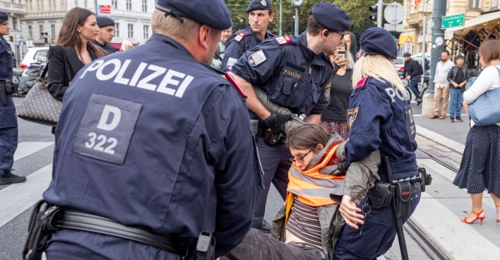 Drei Polizisten tragen eine Aktivistin in einer orangen Warnweste weg.