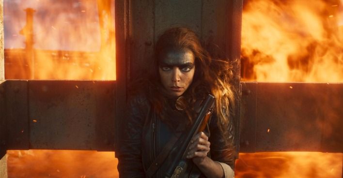 Eine Schauspielerin vom Film "Furiosa: A Mad Max Saga" mit viel schwarzer Schimnke. Hinter ihr sind Flammen. Sie trägt eine Waffe.