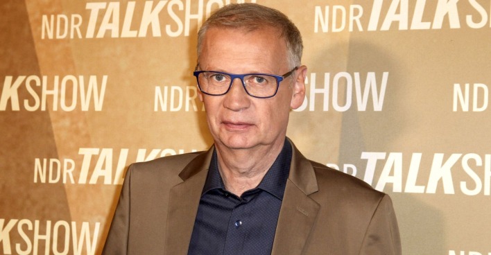 Günther Jauch in mit einem beigen Sakko vor einer beigen Fotowand.