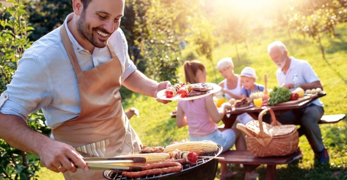 Ein Mann mit schwarzem Vollbart - bekleidet mit einer brauner Schürze grill. In der linken Hand hält er einen Teller mit gegrillten Fleisch und Gemüse, mit der rechten Hand wendet er mithilfe eine Zange einen Maiskolben, der am Grill liegt. Die Sonne scheint, im Hintergrund essen zwei Kinder, eine Frau und ein ältere Herr die gegrillten Speisen.