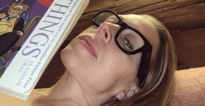 Nina Proll mit Brille räkelt sich auf einem Bett, in der Hand hat sie ein Buch