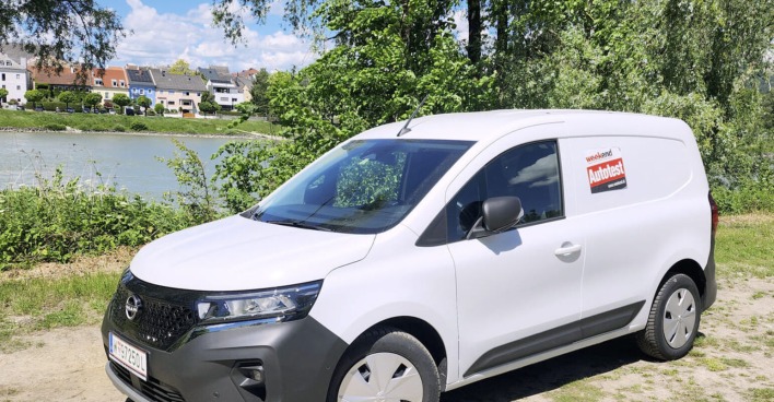 Ein weißer Nissan Townstar glänzt in der Sonne. Das Auto steht am Ufer eines Flusses, auf einem sandigen Parkplatz. Am anderes Ufer sind mehrere Häuser zu sehen.