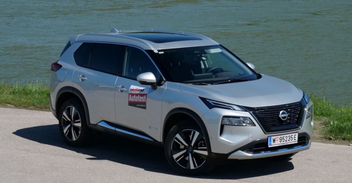 Ein silberner Nissan X-Trail glänzt in der Sonne. Er steht am Ufer eines Flusses auf einem asphaltierten Parkplatz.
