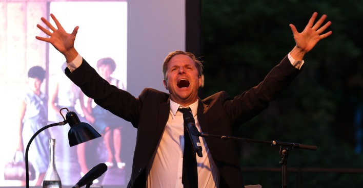 Philipp Hochmair mit einem schwarzen Anzug und einer schwarzen Krawatte auf der Bühne.