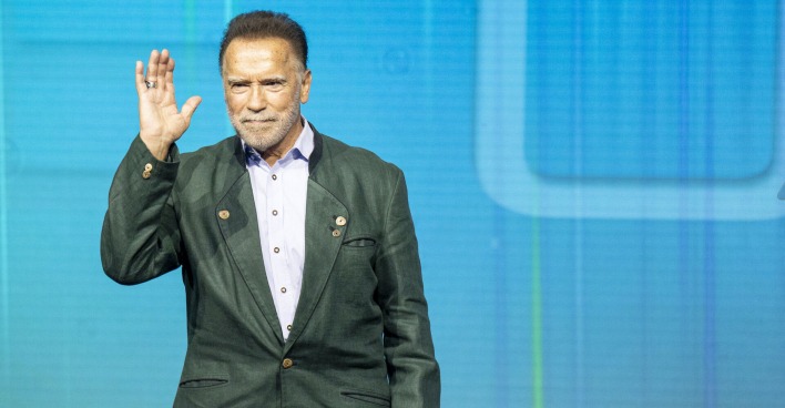 Arnold Schwarzenegger winkt in einem grünen Sakko von der Bühne.