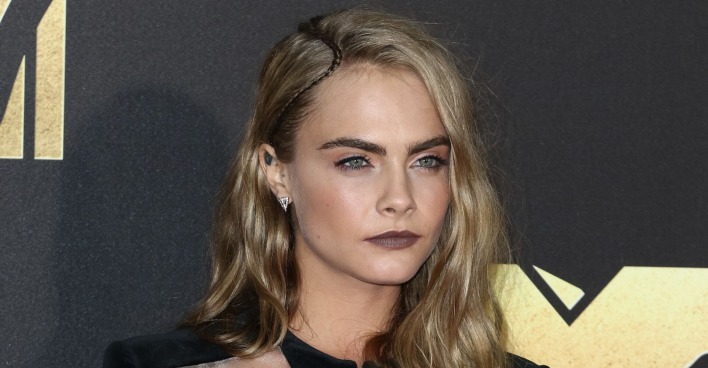 Cara Delevigne mit offenen blonden Haaren und einem dunklen Lippenstift vor einer Fotowand.