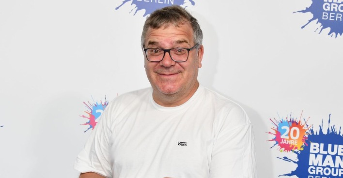 Moderator Elton vor einer Fotowand mit einem weißen T-Shirt.