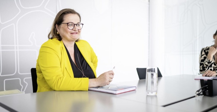 Anette Klinger, Miteigentümerin von Internorm sitzt an einem Schreibtisch auf der ein Notizblock liegt und ein Glas mit Wasser steht. Sie trägt eine braune Brille ein gelbes Sakko und eine schwarze Bluse.