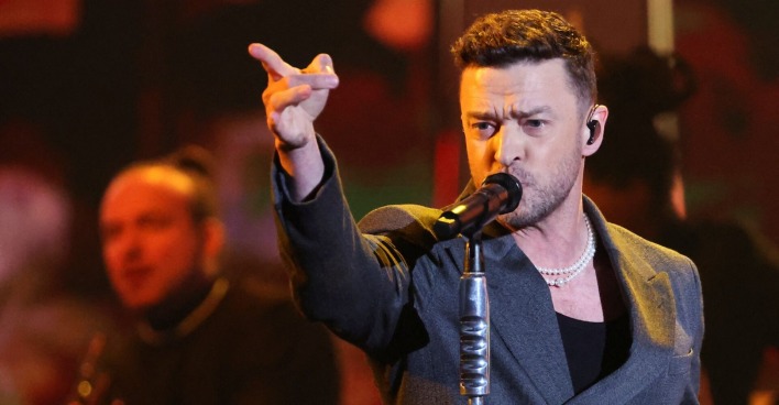 Justin Timberlake mit einem grauen Sakko vor einem Mikrofon. Er zeigt ins Publikum.
