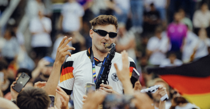 Ein Mann trägt eine Sonnenbrille und spielt ein Saxophon, während ihm alle zujubeln