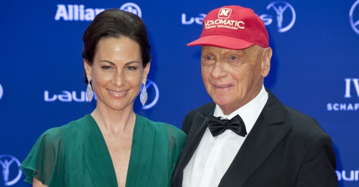 Niki und Birgit Lauda auf einem Event vor einer blauen Fotowand. Sie trägt ein grünes Kleid und Niki Lauda einen Smoking und eine rote Kappe.