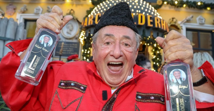 Richard Lugner in rotem Hemd und schwarzem Hut mit zwei Wodkaflaschen in seiner Hand