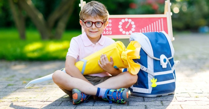 Ein junger Bub - mit kurzer Hose, kurzem Polo und bunten Turnschuhen bekleidet - lacht in die Kamera. Er trägt eine rote Brille und in der Hand hält er eine gelbe Schultüte. Rechts von ihm steht eine blaue Schultasche, hinter ihm eine Kreidetafel auf der Schule zu lesen ist.