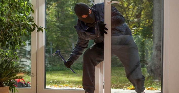 Ein Einbrecher betritt das Haus durch eine Glastüre. Im Hintergrund sind Bäume zu sehen. Der Einbrecher ist dunkel gekleidet, trägt eine Maske und hat schwarze Handschuhe an. In der linken Hand hält er ein blaues Brecheisen.