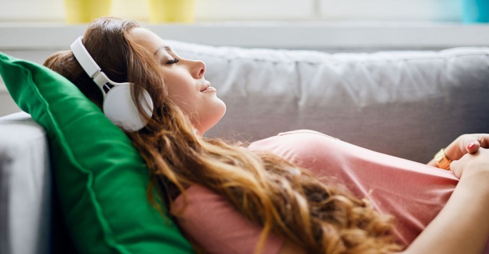 Frau liegt mit Kopfhörern entspannt auf der Couch | Credit: iStock.com/BartekSzewczyk