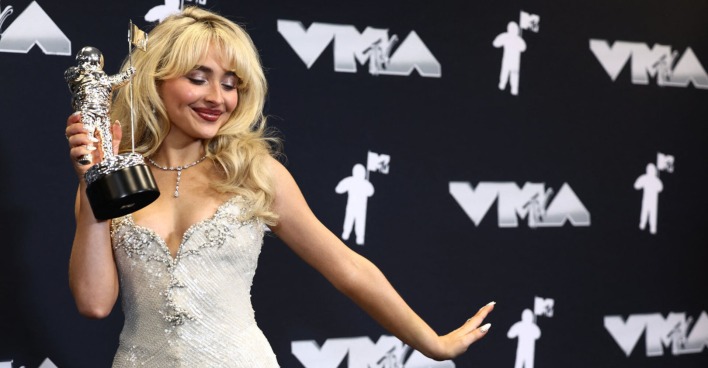 Eine blonde Frau im Glitzerkleid hält eine Trophäe in der Hand