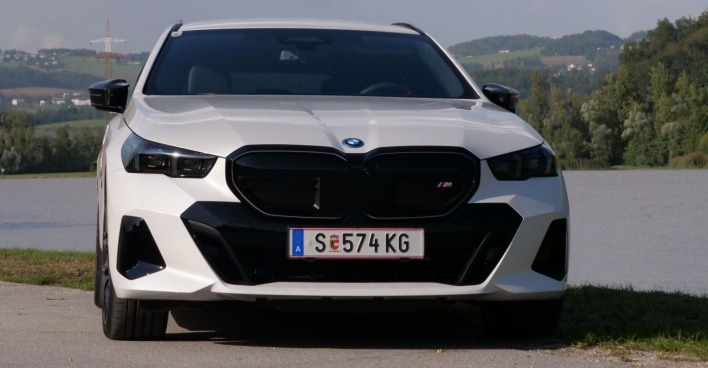 Ein weißer BMW i5 M60 xDrive - mit Salzburger Kennzeichen - steht am Ufer eines Flusses. Da Auto strahlt in der Sonne.