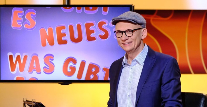 Moderator Baier sitzt vor einem Bildschirm.