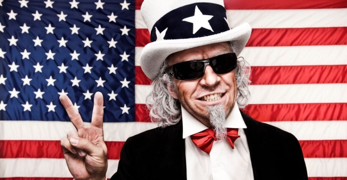 Ein Mann, Symbolfigur Uncle Sam, mit weißem Bart ist zeigt mit seiner rechten Hand das Victory-Zeichen. Er trägt eine schwarze Brille und eine weißen Hut mit schwarzen Band und weißen Sternen. Im Hintergrund ist die Flagge der USA zu sehen.
