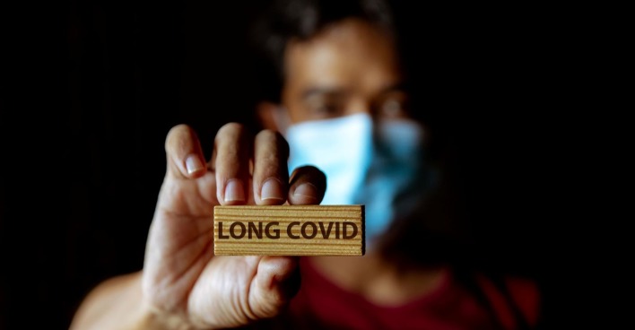 Mann trägt Corona-Maske und hält Holzstück mit Aufschrift "Long Covid" in die Kamera