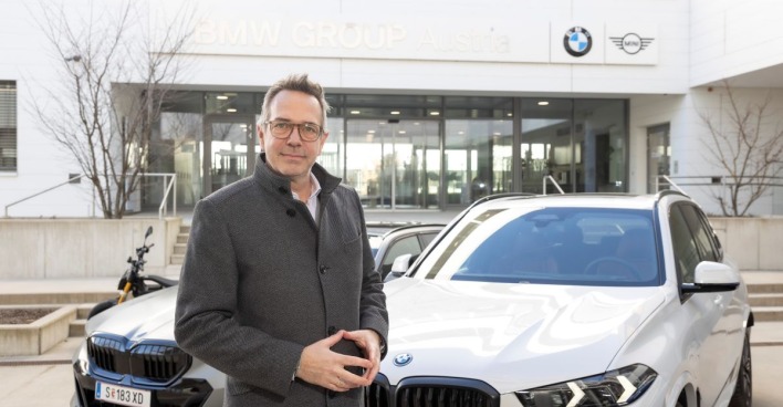 Alexander Bamberger, trägt eine Brille und ist mit einem grauen Mantel bekleidet. Der BMW-Manager steht vor zwei weißen BMWs. Im Hintergrund ist ein Gebäude mit einer großen Glasfläche zu sehen. Am Gebäude ist das BMW-Logo angebracht.