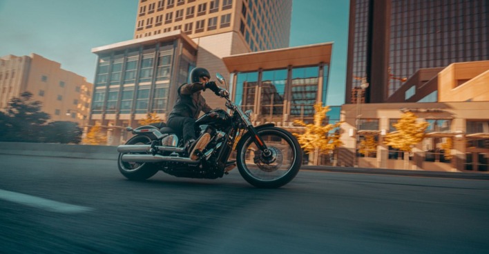 Ein Biker mit Helm sitzt auf einer Harley-Davidson. Er fährt im Zentrum einer Großstadt, im Hintergrund sind Hochhäuser zu sehen. Die Bäume im Hintergrund tragen herbstlich gefärbte Blätter, die in der Sonne gelb leuchten.