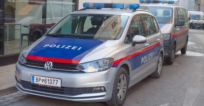 Zwei österreichische Polizeiautos, ein silberner VW mit der Aufschrift "Polizei" in Blau und Rot sowie ein weiteres Einsatzfahrzeug, parken auf einer Straße in der Grazer Innenstadt. Im Hintergrund sind Gebäude und ein Schaufenster zu sehen.