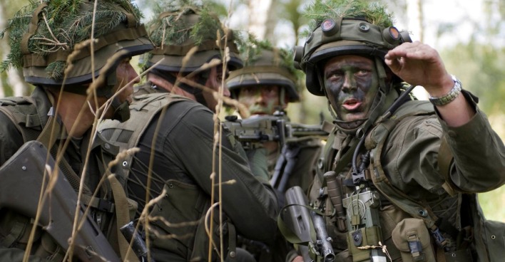 Soldaten des österreichischen Bundesheeres in voller Ausrüstung während einer Übung im Wald. Die Gesichter sind mit Tarnfarbe bemalt, Helme mit Zweigen getarnt. Ein Soldat gibt gestikulierend Anweisungen