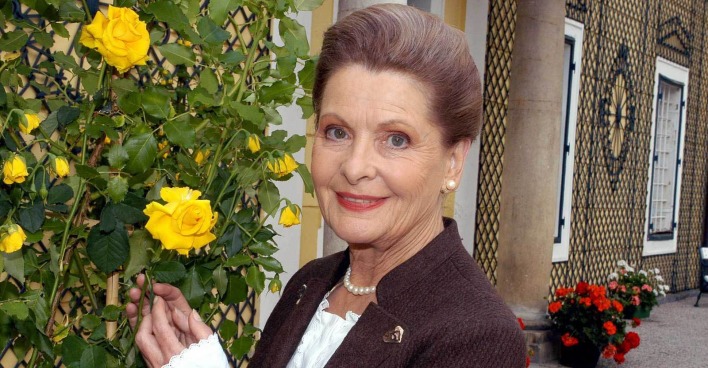 Johanna Matz steht lächelnd neben einem blühenden Rosenstrauch mit gelben Rosen. Sie trägt ein braunes Kostüm mit Perlenkette und blickt freundlich in die Kamera. Im Hintergrund ist ein Gebäude mit kunstvoll verzierten Fenstern und Blumenschmuck zu sehen.