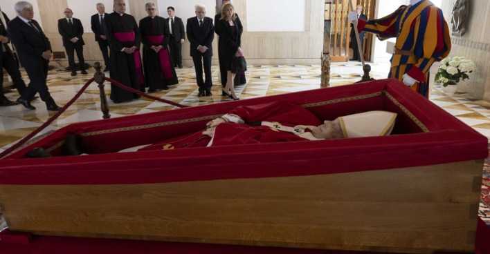 Papst Franziskus liegt im offenen Sarg in rotem liturgischem Gewand. Um den Sarg versammeln sich Geistliche, Politiker und ein Mitglied der Schweizergarde.