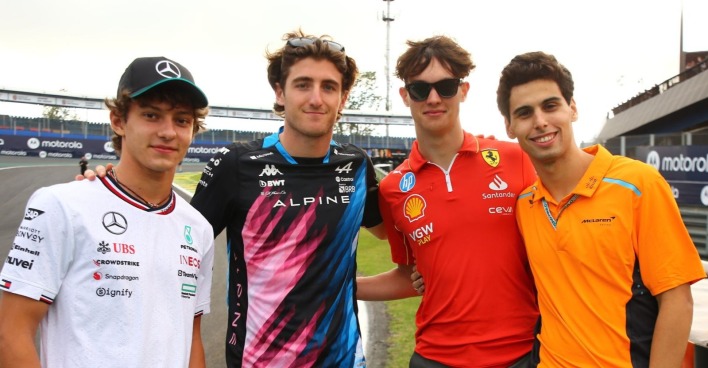 Vier junge Formel-1-Fahrer in Teamkleidung von Mercedes, Alpine, Ferrari und McLaren posieren gemeinsam an der Strecke.