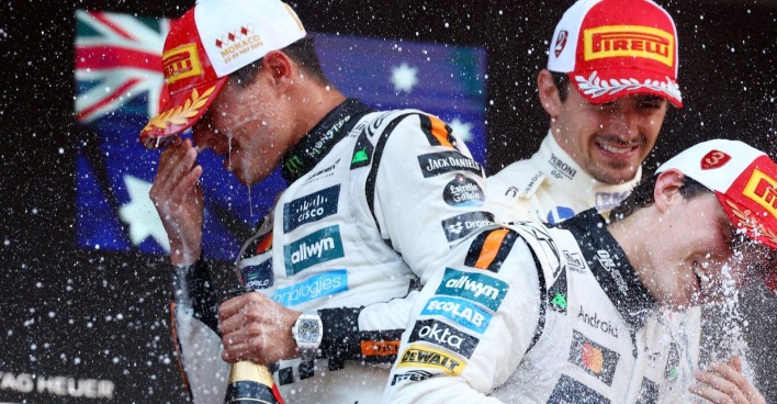 Lando Norris spritzt mit Champagner, neben ihm Oscar Piastri und Charles Leclerc auf dem Podium des Formel-1-Rennens in Monaco 2025, alle tragen Pirelli-Kappen und Rennanzüge.