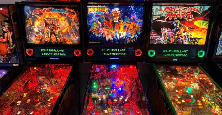 Drei beleuchtete Flipperautomaten nebeneinander in einer Spielhalle. Die Titel der Maschinen sind von links nach rechts: "Medieval Madness", "Monster Bash" und "Attack from Mars". Die Spielflächen sind bunt beleuchtet und zeigen jeweils thematisch passende Designs und Figuren.