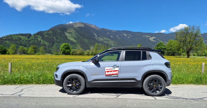 Ein hellblauer Jeep Avenger parkt auf einer Straße. Im Hintergrund sind eine Wiese mit gelben Blumen zu sehen und einige Obstbäume. Dahinter sieht man auch - nicht sehr hohe - Berge - die bewaldet sind.