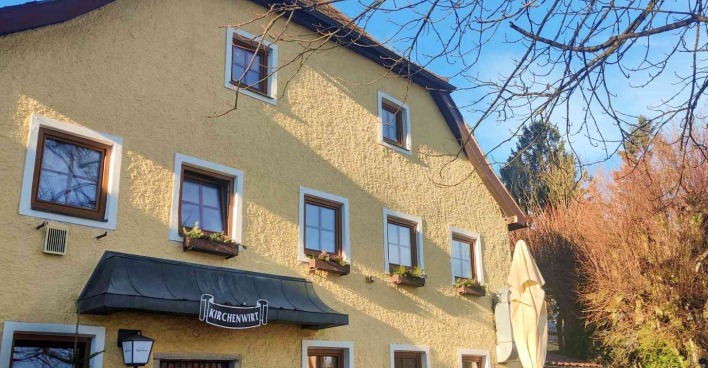 Gelbes, traditionelles Gasthaus mit dem Namen „Kirchenwirt“ auf einem Schild über dem Eingang. Das Gebäude hat mehrere Fenster mit Blumenkästen und ist von kahlen Bäumen sowie herbstlich gefärbtem Gehölz umgeben. Der Himmel ist klar und blau.