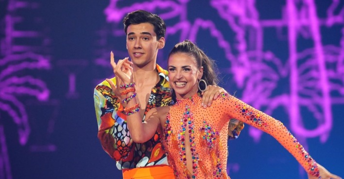 Diego Pooth und seine Tanzpartnerin Ekaterina Leonova lächeln auf der "Let's Dance"-Bühne im Finale 2025. Beide tragen bunte, glitzernde Kostüme, die Kulisse ist violett beleuchtet.