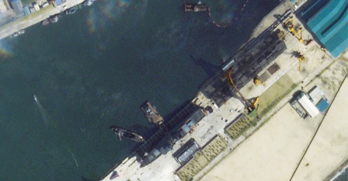Satellitenbild eines nordkoreanischen Werftgeländes in Chongjin. Ein großes Kriegsschiff liegt seitlich im Wasser, umgeben von Kränen und Werftbauten entlang der Kaimauer.