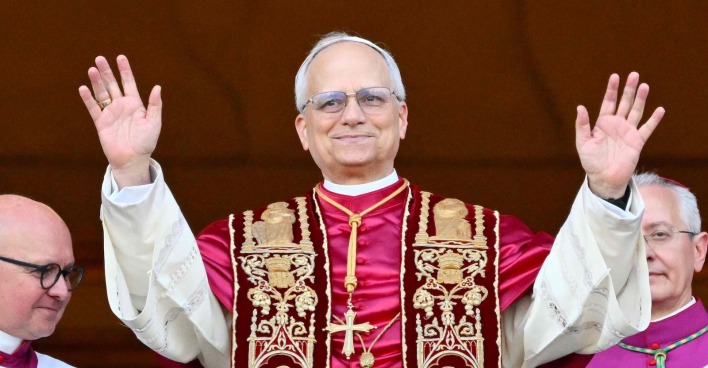 Papst Leo XIV. steht in päpstlicher liturgischer Kleidung auf dem Balkon des Petersdoms und hebt beide Hände zum Gruß. Links und rechts neben ihm stehen Kirchenvertreter.