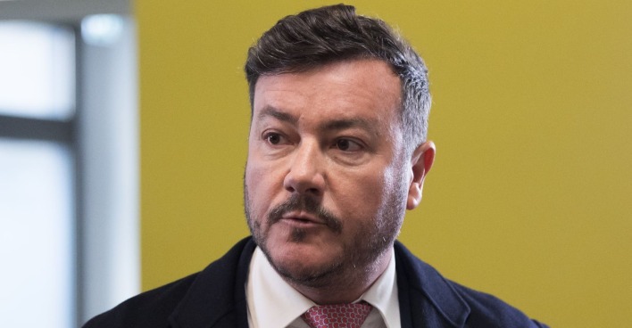 René Benko blickt ernst zur Seite, im Hintergrund eine gelbe Wand.