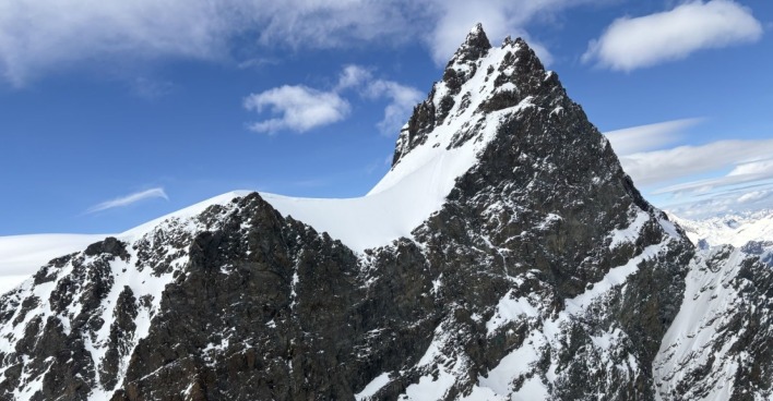 Verschneiter, steil aufragender Gipfel des Rimpfischhorns in den Walliser Alpen unter blauem Himmel mit vereinzelten Wolken. Links davon ein schneebedeckter Sattel, rechts steile Felsflanken.