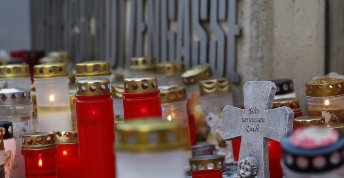 Zahlreiche rote und weiße Grablichter stehen vor einer Wand mit dem Schriftzug „Dreierschützengasse“. Im Vordergrund ein graues Steinkreuz mit der Inschrift „Wir vermissen Dich“ und einer Engelsfigur.