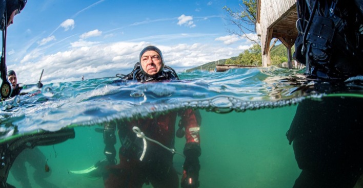 Taucher in voller Ausrüstung im Attersee, teilweise unter Wasser fotografiert, mit Blick auf einen Holzsteg und das Ufer im Hintergrund.