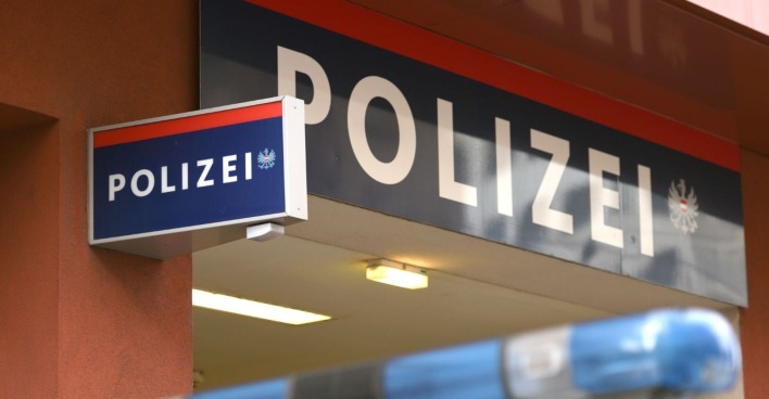 Eingangsbereich einer Polizeiinspektion in Österreich mit blau-rotem Polizeischriftzug und Blaulicht eines Polizeiwagens im Vordergrund.