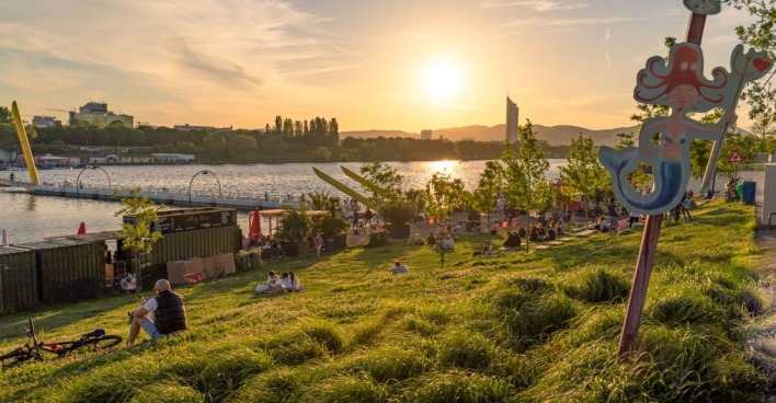 alte Donau im Sommer, Menschen sitzen auf Wiese am Wasser und beobachten den Sonnenuntergang