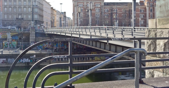 Blick auf die Augartenbrücke in Wien mit Blick auf den Donaukanal und Graffiti an den Ufermauern; im Vordergrund ein Geländer mit Schmierereien, im Hintergrund historische Gebäude.