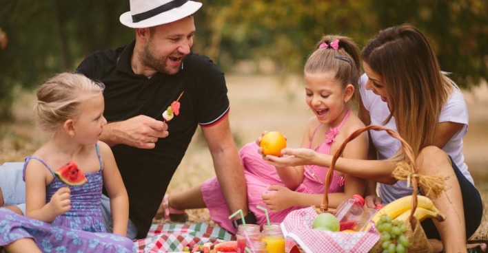 Eine fröhliche Familie beim Picknick im Freien. Zwei Kinder und ihre Eltern sitzen auf einer Decke, essen frisches Obst und lachen miteinander. Der Vater trägt einen weißen Hut und hält einen Obstspieß, während die Mutter und eine Tochter eine Orange betrachten. Im Vordergrund steht ein Picknickkorb mit Bananen, Trauben und Getränken. Die Szene findet an einem sonnigen Tag im Grünen statt.
