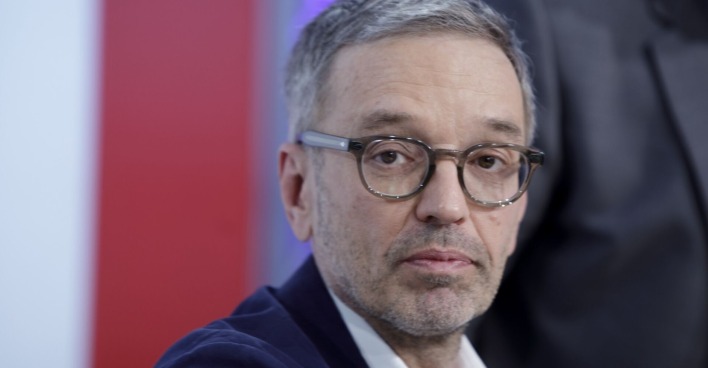 Herbert Kickl mit ernstem Gesichtsausdruck, trägt Brille und Sakko, vor rotem und weißem Hintergrund.